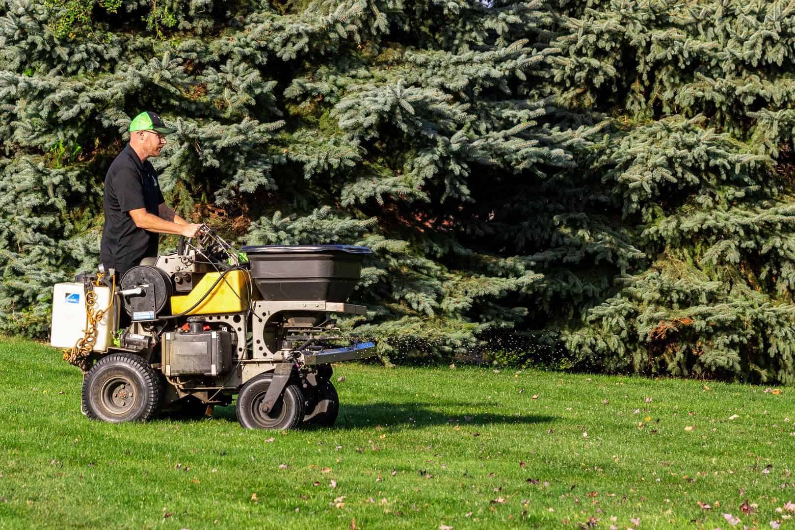 An Ecolawn technician applying fall fertilizer with a ride-on spreader across a green lawn